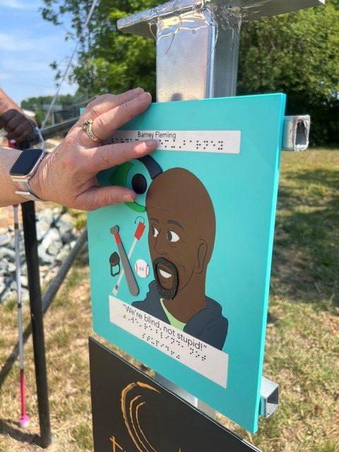 A hand rests on a teal tactile trail marker honoring Barney Fleming. The marker shows his name in raised print and braille, an illustrated portrait, tactile icons of a long cane and an audio device, and the quote 