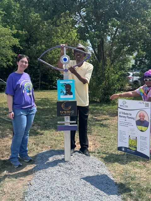 A smiling man in a yellow shirt and hat reaches up to touch a tall outdoor sculpture shaped like a stylized eye with a blue iris, mounted on a pedestal along the trail, while two other visitors look on.