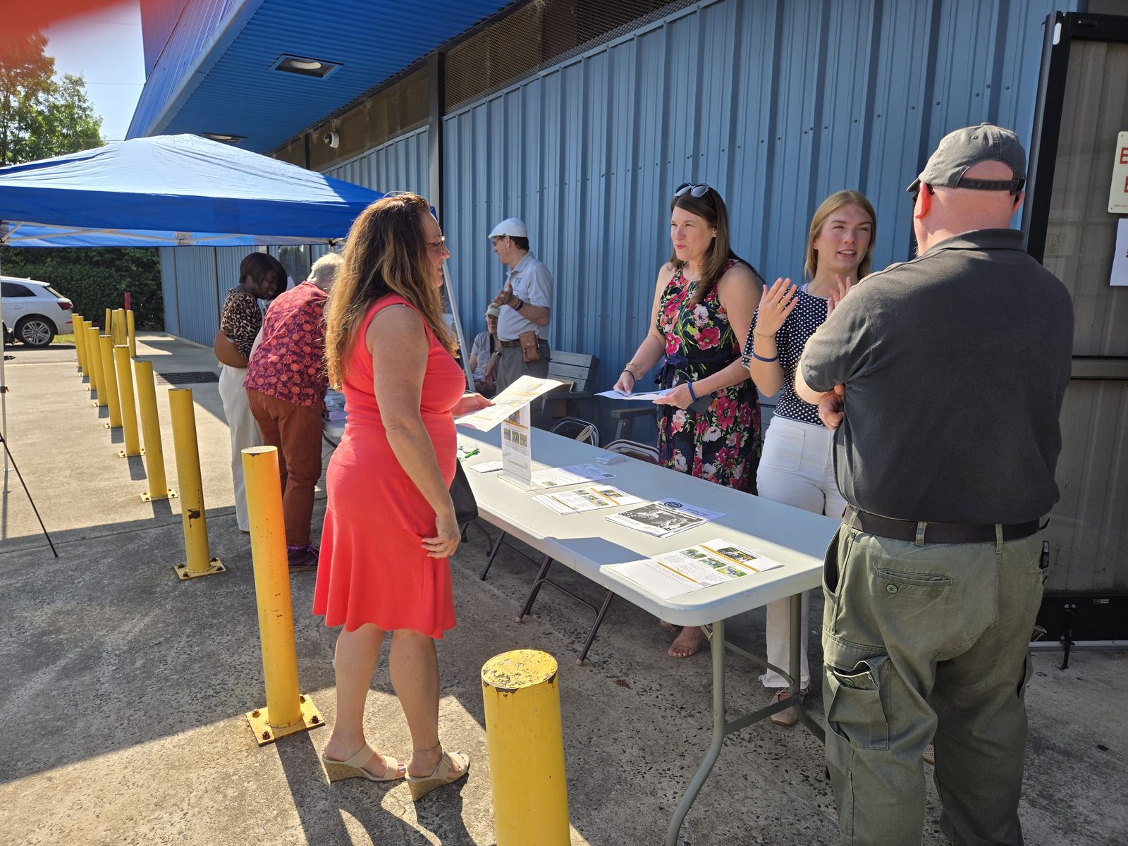 Two hosts greet a guest at an outdoor welcome table covered with printed programs and information about the Charlotte Braille Trail.