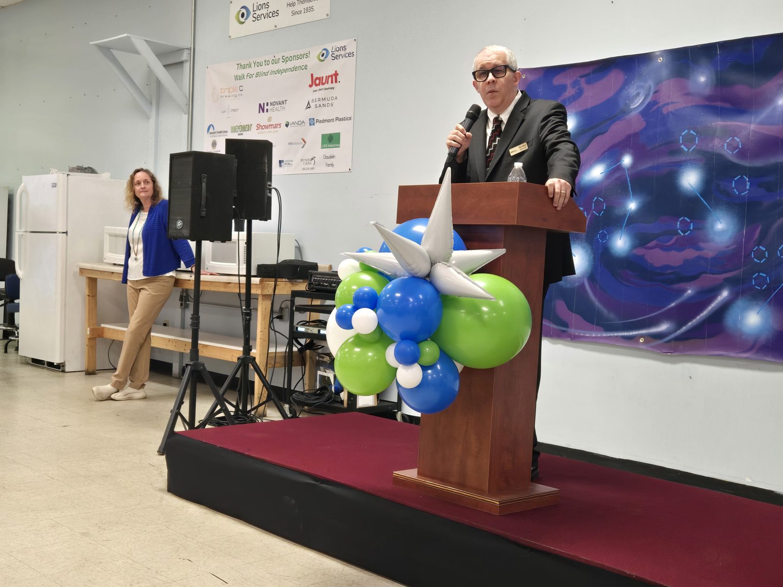 A speaker in a dark suit addresses guests from a wooden podium decorated with blue, green, and white balloons, in front of a 