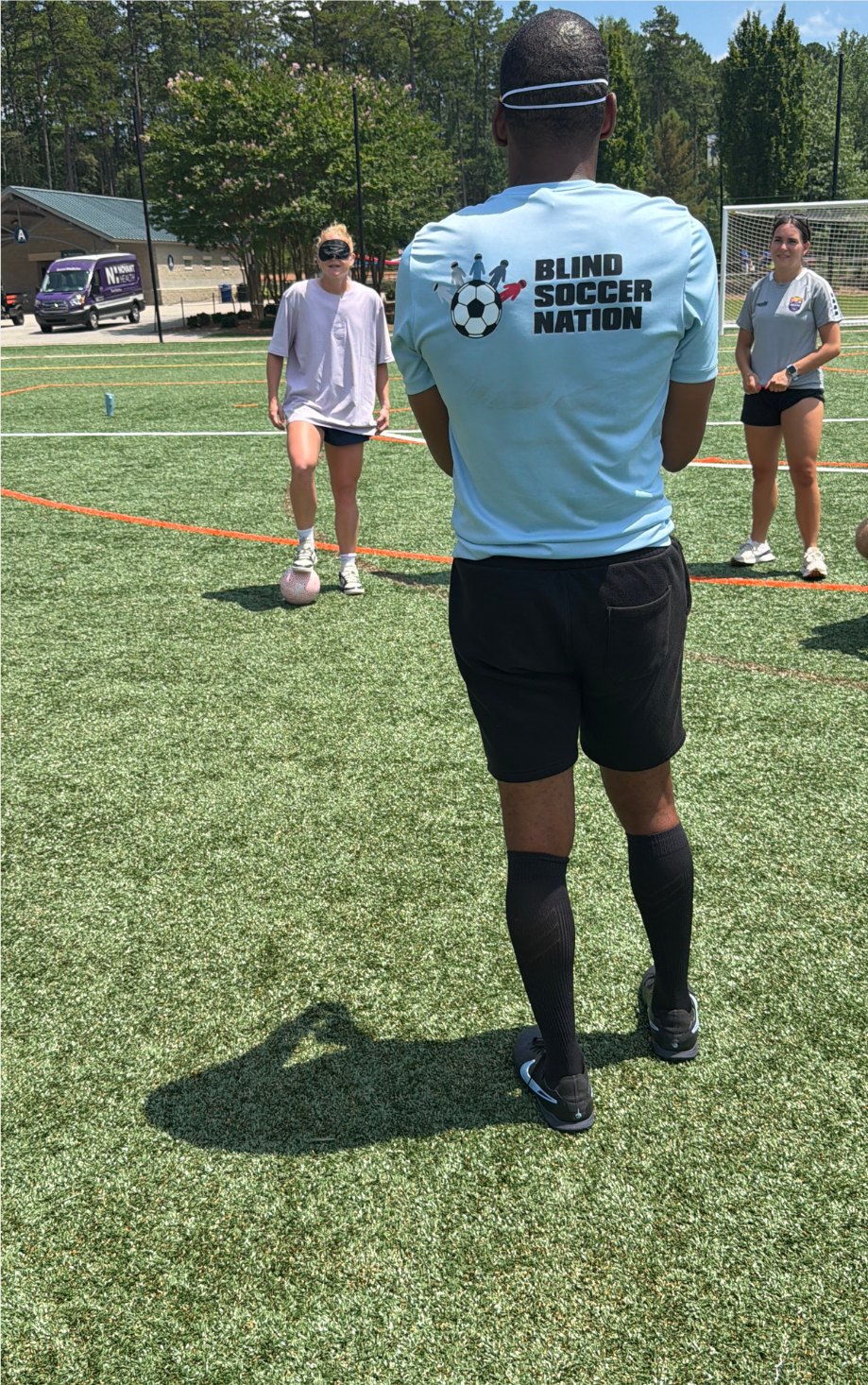 From behind, a Blind Soccer Nation coach in a light blue shirt faces players on a green turf field under a sunny sky.