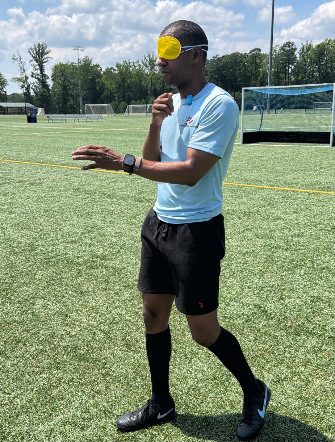 Antoine Craig, wearing a yellow eyeshade and light blue Blind Soccer Nation kit, claps while coaching on the turf field.