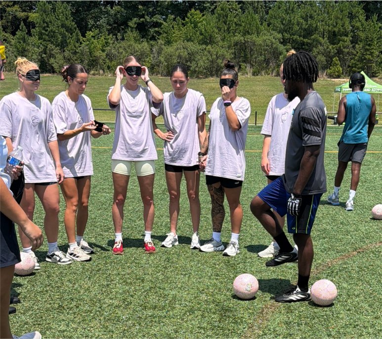 Five participants in white shirts stand in a circle wearing eyeshades while a coach demonstrates with soccer balls at their feet.