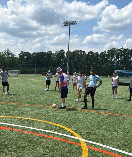 Players and coaches gather near the sideline on a sunny turf field at the Blind Soccer Expo.